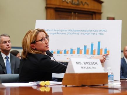 A woman sitting at a table with a microphone in front of her. She is wearing a black blazer and glasses and appears to be giving a presentation. On the table there is a large white board with a bar graph and text that reads "Auto-Injector Wholesale Acquisition Cost (WAC) Average Revenue (Net Price) to dylan Per 2-Pack". There are other people sitting in the background but they are not visible. The woman is pointing to the graph and seems to be explaining it to the audience.