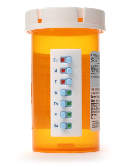 Of an orange prescription bottle with a white lid. The bottle is cylindrical in shape and has a label on the right side. The label has a white background with black text. On the left side of the bottle there is a white remote control with blue and green buttons. The buttons are arranged in a grid-like pattern. The remote control appears to be used to control the contents of the prescription bottle.