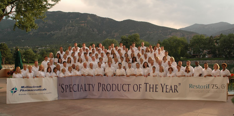 A large group of people standing in front of a large banner that reads "Specialty Product of the Year" and "Restoril 75". The group is made up of both men and women all wearing white lab coats. They are arranged in a semi-circle facing the camera. In the background there are mountains and trees and the sky is blue with a few clouds. The group appears to be posing for a photo and there is a table and chairs on the right side of the image.