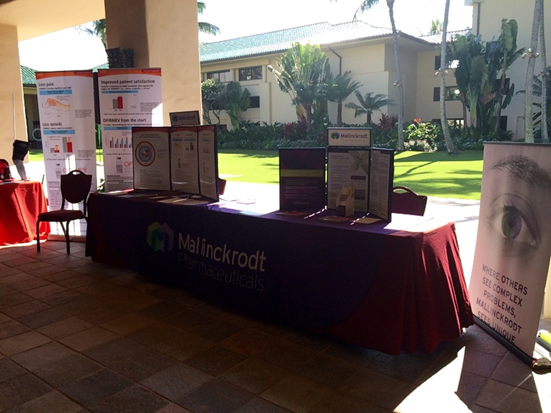 A table with a red tablecloth and a banner that reads "Mallinckrodt Pharmaceuticals". On the table there are several informational boards with information about the company's products and services. The table is set up in a room with a tiled floor and a large window in the background. There are palm trees and a building visible in the distance. On the right side of the image there is a banner with an image of an eye and the company logo.