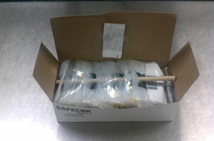 A cardboard box with the word "SAFECOR" written on it. Inside the box there are several items that appear to be medical equipment. The items are wrapped in plastic and are arranged neatly in the box. There is a white label on the top right corner of the box with a barcode and some text. The box is sitting on a metal surface.