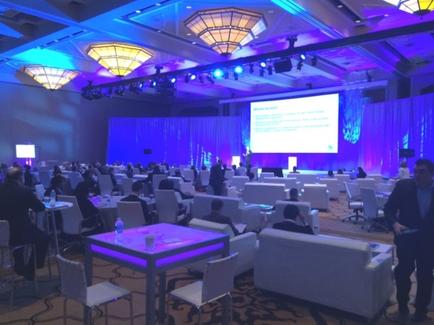 A large conference room with a high ceiling and multiple chandeliers hanging from it. The room is filled with white sofas and chairs arranged in rows with a stage in the center. On the stage there is a large screen displaying a presentation. There are several people sitting at tables and chairs attentively listening to the presentation. The walls are decorated with blue and purple lights creating a warm and inviting atmosphere. The overall mood of the room is professional and professional.