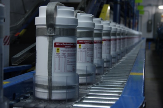 A row of white plastic containers with labels on them arranged on a conveyor belt in a factory. The containers appear to be cylindrical in shape and have a handle on top for easy carrying. The labels on the containers are red and white with the brand name "Ultra-Tech" written in bold letters at the top. The background is blurred but it appears to be a large industrial space with other machines and equipment visible.