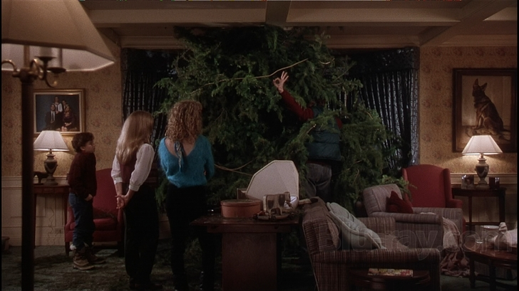 A living room with a large Christmas tree in the center. The tree is decorated with colorful ornaments and lights. There are three people in the room two women and a child standing around the tree. The woman on the left is wearing a blue sweater and the woman in the middle is wearing an orange sweater. The child on the right is standing next to the tree and appears to be looking at it. The room is furnished with a red armchair a coffee table and a lamp. The walls are painted in a warm yellow color and there are several framed pictures hanging on the walls. The floor is covered with a patterned rug.