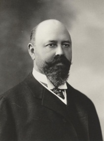 A black and white portrait of a man. He is a middle-aged man with a bald head and a full beard. He has a serious expression on his face and is looking directly at the camera. He appears to be in his late twenties or early thirties. The man is wearing a suit and tie with a collared shirt underneath. The background is plain and blurred putting the focus on the man's face. The photograph is in a studio setting with no other people or objects in the frame.