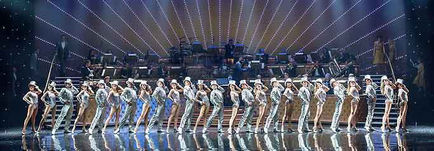 A group of dancers performing on a stage. The dancers are arranged in a line with their arms and legs stretched out in front of them. They are all wearing white outfits with gold accents. The stage is lit with bright lights and there is a large screen in the background. The audience can be seen in the back watching the performance. The overall mood of the image is energetic and dramatic.