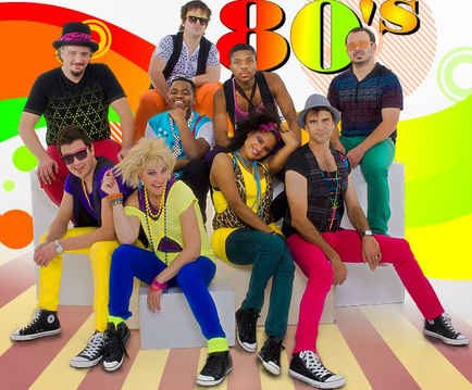 A group photo of a group of young people dressed up in 80s style clothing. There are nine people in the photo six men and three women all wearing colorful outfits and accessories. They are sitting on white cubes and posing for the camera. The background is a colorful abstract design with the word "80's" written in bold orange letters at the top. The overall mood of the photo is vibrant and energetic.