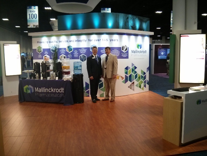 Two men standing in front of a booth at a trade show. The booth is decorated with a blue and white color scheme and has a large banner that reads "Mallinckrodt Pharmaceuticals" in bold letters. The banner also has images of various pharmaceutical products and a logo for the company. The men are wearing suits and are posing for the photo. On the right side of the booth there is a desk with a computer monitor and other equipment. In the background there are other booths and a large screen displaying information about the company's products. The floor is made of wood and there are banners hanging from the ceiling.