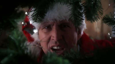 A still from a movie or TV show. It shows a close-up of a man's face who appears to be in his late 40s or early 50s wearing a white Santa hat with a red pom-pom on top. He is looking directly at the camera with a surprised or shocked expression on his face. The background is blurred but it seems like he is standing in front of a Christmas tree with ornaments hanging from it. The overall mood of the image is one of excitement or surprise.