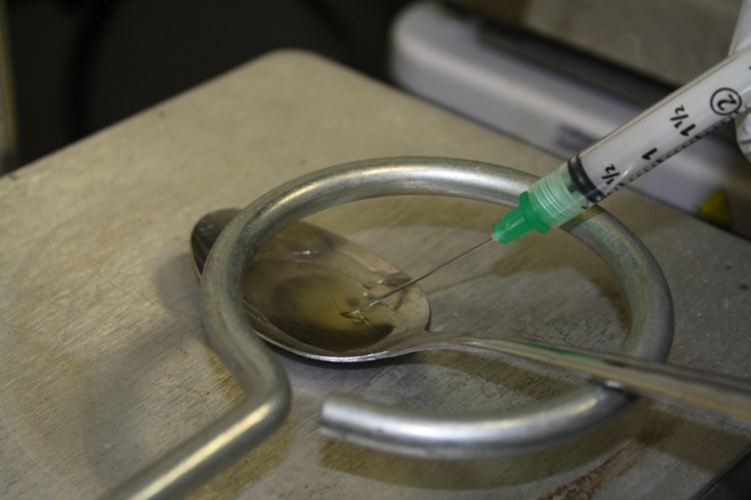 A close-up of a medical instrument specifically a syringe with a green liquid being injected into a metal spoon. The syringe is being held in place by a person's hand and the spoon is resting on a metal surface. The spoon appears to be made of stainless steel and has a curved handle. The background is blurred but it seems to be a laboratory setting.