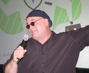 A man standing on a stage with a microphone in his hand. He is wearing a black button-down shirt and a blue beanie. He has a pair of sunglasses on and is smiling as he speaks into the microphone. Behind him there is a green wall with a logo that reads "2007". The man appears to be giving a speech or presentation.