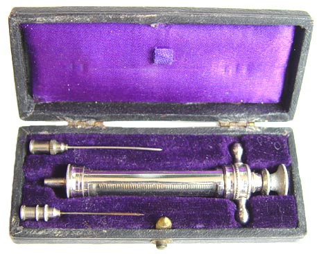 An antique medical instrument specifically a syringe in a black velvet case. The syringe is made of metal and has a long cylindrical body with a pointed tip and a handle on one end. The handle is decorated with a small knob on the top and a small ball at the bottom. The case is open revealing the syringe inside. The inside of the case is lined with a purple velvet lining and there is a small purple velvet pouch on the right side.