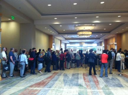 A large group of people standing in a long line in a lobby. The lobby has a high ceiling with recessed lighting and a chandelier hanging from it. The people are of different ages genders and ethnicity's and they are all looking towards the right side of the image. They appear to be waiting in line with some people holding signs or banners. The floor is covered in a patterned carpet and there are pillars on either side. The overall atmosphere of the lobby is busy and bustling.