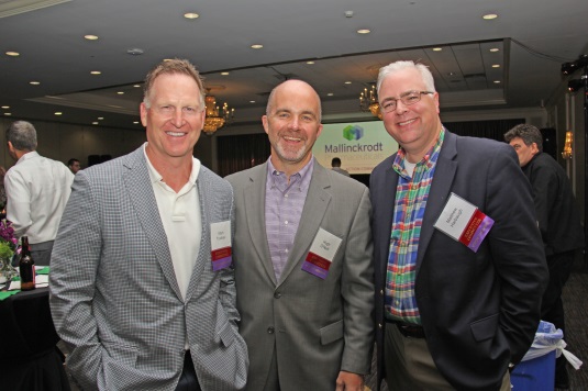 Three men standing together and posing for a photo. They are all smiling and appear to be at an event. The man on the left is wearing a gray suit and has short blonde hair while the man in the middle has a bald head and is balding. All three men are wearing name tags around their necks. The background shows a large room with a chandelier and other people in the background. It appears to be a conference or event space with tables and chairs set up.