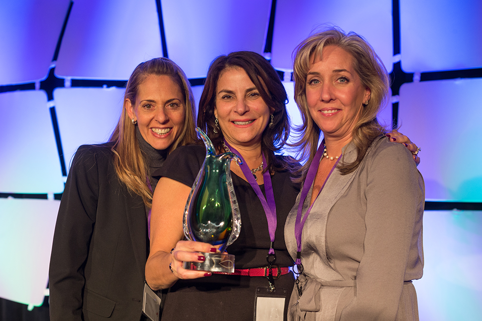 Three women standing on a stage with a blue and white background. The woman in the middle is holding a large green trophy with a red ribbon around it. She is wearing a black dress and has a name tag around her neck. On the left is a blonde woman with long hair and on the right is a brunette woman with shoulder-length hair. All three women are smiling and appear to be posing for the photo. They are standing close together and are standing in front of a large screen with a geometric pattern.