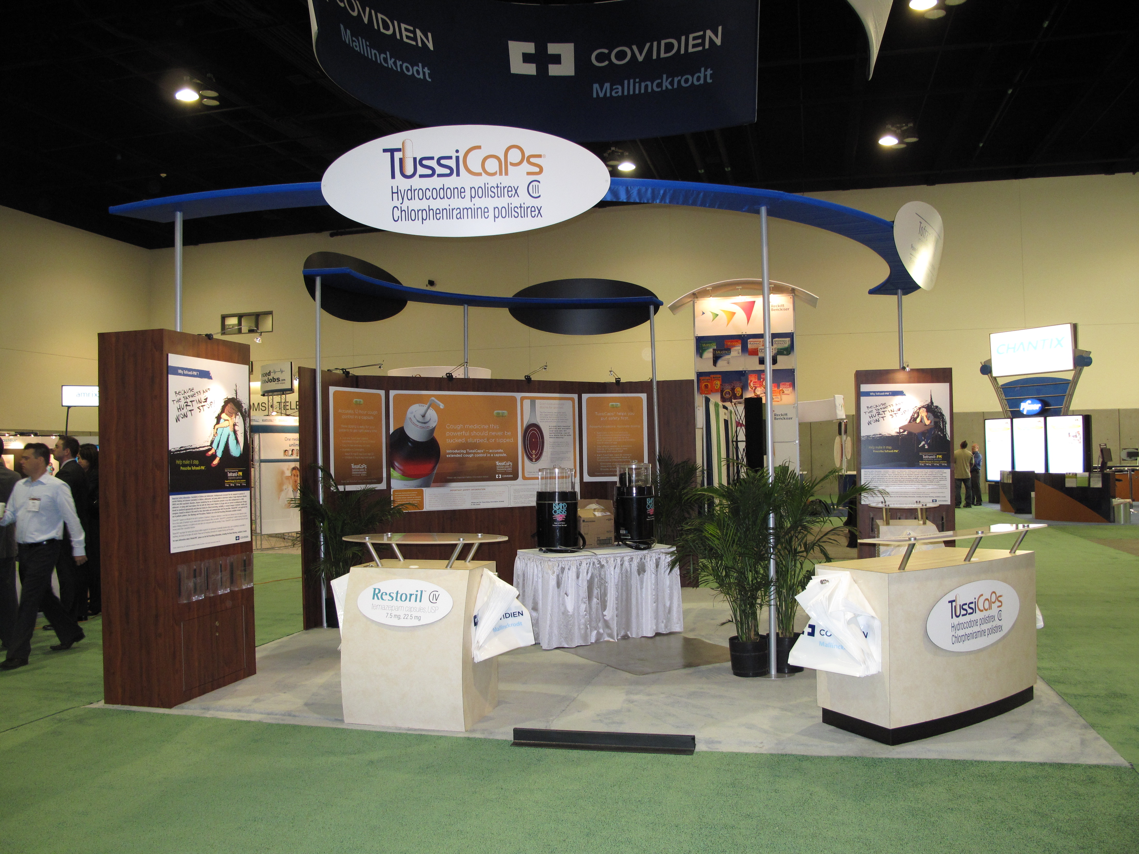 A trade show booth with a blue and white color scheme. The booth has a large banner that reads "TussiCaps" and "Covidien" in bold letters. Below the banner there is a sign that says "Mallinckrodt" in smaller letters. <br /><br />The booth is set up in a large hall with a green carpeted floor and white walls. There are several people walking around the booth some of whom are wearing business attire. On the left side of the image there are two large banners with information about the company's products and services.<br /><br />In the center of the booth is a table with a white tablecloth and a plant in front of it. There is also a large screen on the right side which appears to be displaying information about a product or service. The overall atmosphere of the exhibition hall is modern and professional.