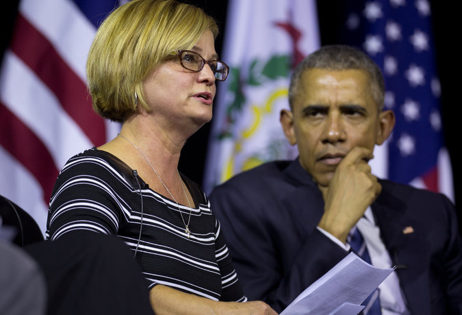 Former US President Barack Obama and a woman sitting side by side in front of two flags. The woman is on the left side of the image wearing a black and white striped shirt and glasses. She appears to be speaking with her hand resting on her chin and a serious expression on her face. The man on the right side is wearing a suit and tie and is looking off to the side. The flags in the background are the American flag and the flag of the United States. The image appears to have been taken at a press conference or event.