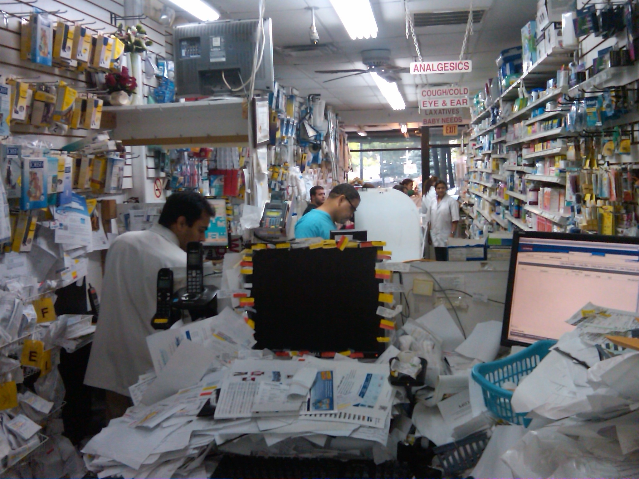 The interior of a pharmacy store. The store is filled with shelves stocked with various medicines and products. In the foreground there is a counter covered with papers computer monitors and cordless telephones. In the midground employees in white lab coats - perhaps pharmacists? - stand near ready to assist customers. Multiple customers wait for service in the background spaced out in narrow aisles. The store appears to have a cluttered and disorganized appearance.