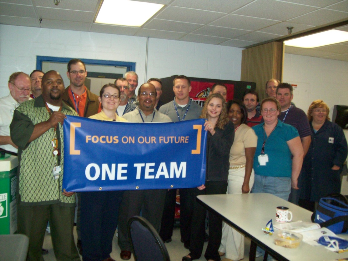 A group of people standing in a room with a large blue banner that reads "Focus on our future one team". The group is composed of men and women of different ages and ethnicities and they are all smiling and posing for the photo. Some of them are holding the banner with their hands while others are standing around a table with various items on it. The room appears to be an office or conference room with white walls and a desk in the background.