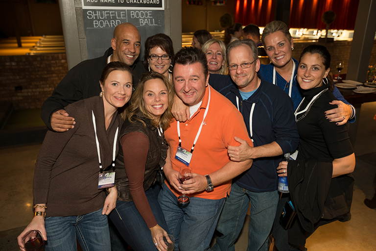 A group of nine people posing for a photo in a restaurant. All are smiling and looking at the camera. They are standing close together and appear to be happy and relaxed. In the background there is a sign that reads "Add name to chalkboard" for shuffleboard and bocce. In the background there is a table with plates and glasses on it. The restaurant appears to be dimly lit with red curtains.