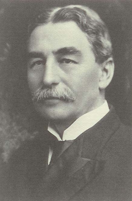 A black and white portrait of a man. He appears to be in his late twenties or early thirties with short wavy hair and a mustache. He is wearing a suit and tie and is looking directly at the camera with a serious expression on his face. The background is blurred so the focus is on the man's face.