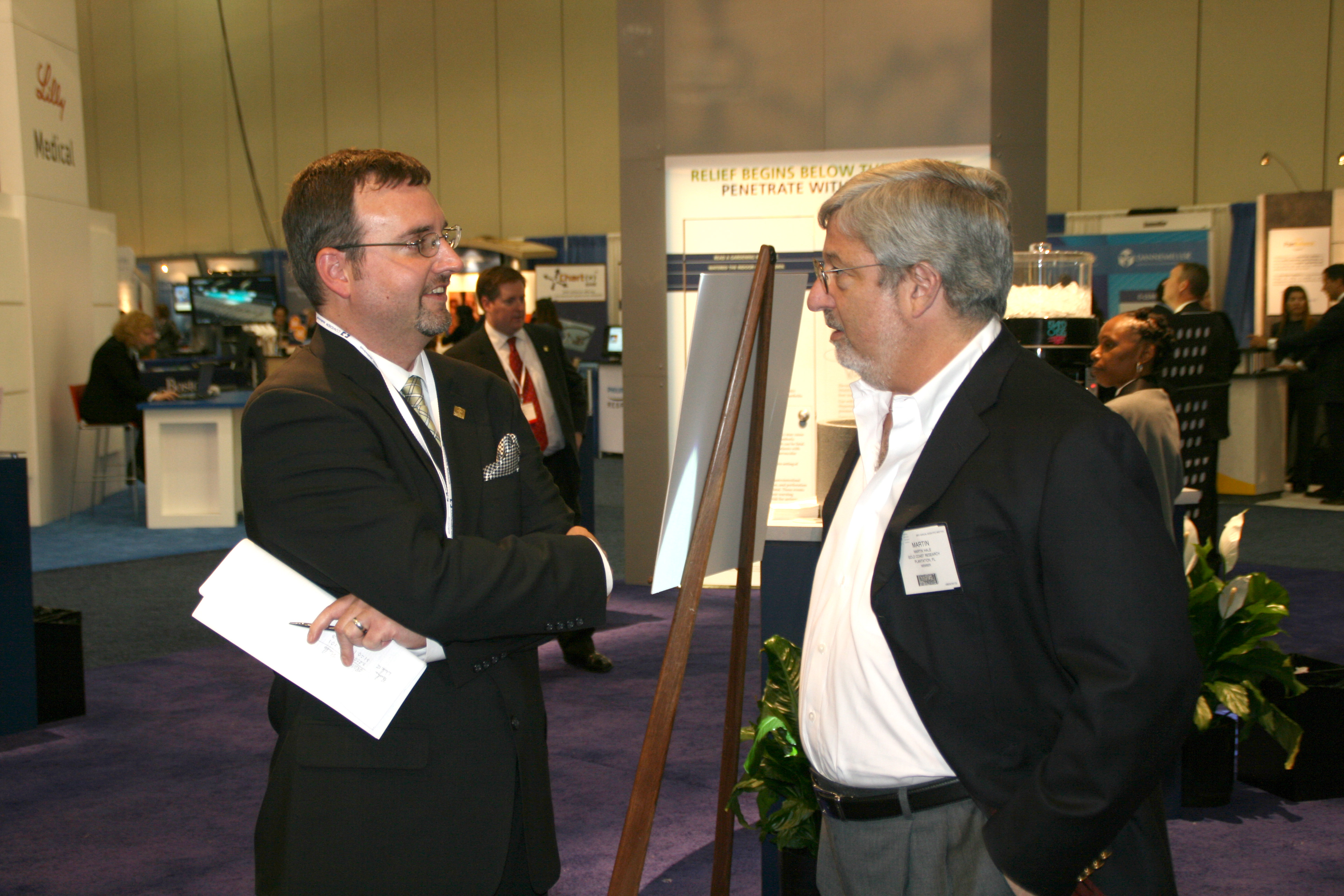 Two men engaged in a conversation at an exhibition. They are standing in a large room with a purple carpet and white walls. The man on the left is wearing a black suit and glasses and is holding a piece of paper in his hand. He appears to be explaining something to the other man who is also wearing a suit and has a name tag on his shirt. There is a sign on a easel in front of them that reads "Welcome to the exhibition". In the background there are other people and booths with information about the exhibition.