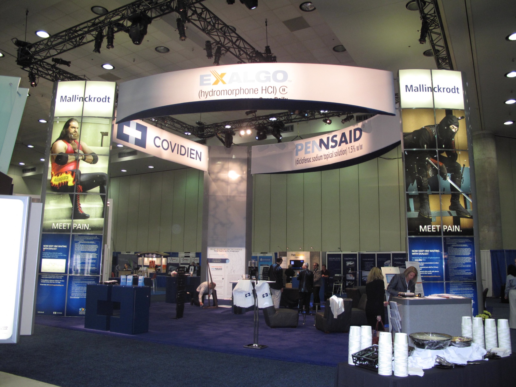 A trade show booth with a large banner that reads "Exalgo" and "Covidien" on both sides. The booth is located in a large hall with a high ceiling and large windows. There are several people gathered around the booth some standing and some sitting at tables. On the left side of the image there is a large screen displaying the company's logo and contact information. In the center of the booth is a sign that says "Pennsaid" with a picture of a man in a suit and tie. The floor is covered with a blue carpet and there are several tables and chairs scattered around the room.