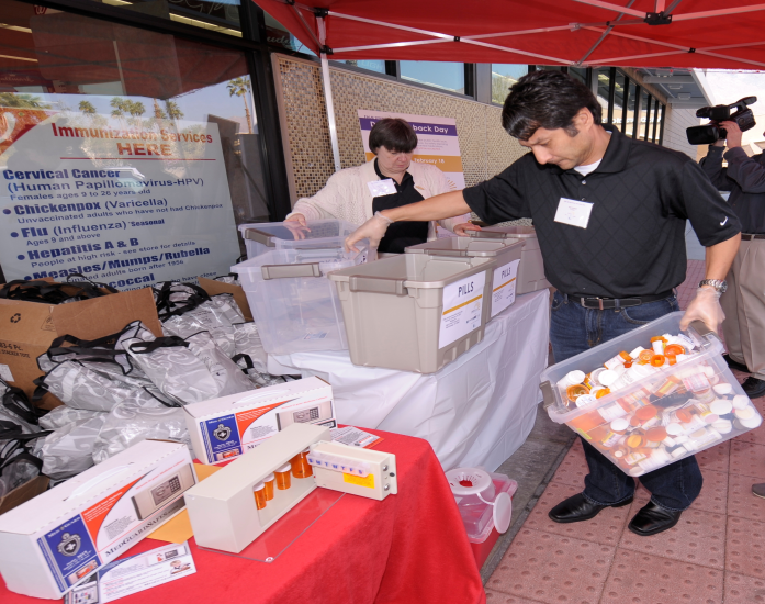 Two people working at a medication disposal event with a cameraman in the background. A man in business casual clothing is carrying a clear plastic tote full of prescription pill bottles while examining an opaque plastic tote labeled "Pills" on its front. A woman in business clothing is examining multiple plastic totes perhaps as part of sorting medications. Promotional items (including tote bags) and paraphernalia (ex : a medication lock box) are visible in the foreground. The event appears to be taking place outside near an office building under a red portable awning.