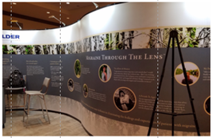 A curved wall with a large banner on it. The banner has multiple images and text on it including a photo of a person's face a camera and a tripod stand. The text on the banner reads "Explore through the lens" and there is a logo of the company "older" on the top left corner. The wall appears to be in a conference room or exhibition space as there are chairs and a table visible in the background.