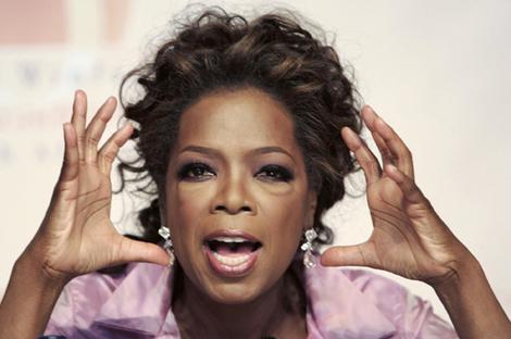 A close-up of a woman's face. She appears to be in her late 40s or early 50s with dark hair styled in loose curls. She is wearing a pink blouse and has a surprised or shocked expression on her face. Her hands are raised above her head as if she is making a point or making a statement. The background is blurred but it seems like she is at a press conference or interview.