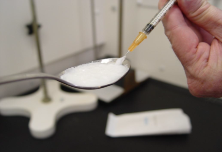 A spoon filled with a white substance likely heroin or another drug and a hand holding a syringe. The syringe drawing up the substance.  The background is blurred but it appears to be  laboratory setting with white cabinets and a black countertop.