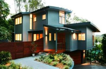 A modern two-story house with a unique design. The house is located in a wooded area with trees and shrubs surrounding it. The exterior of the house is painted in a dark grey color with large windows on both floors and a sloping roof. The front entrance has a wooden staircase leading up to the second floor with a wooden railing on either side. There is a small garden in front of the entrance with a variety of plants and trees. The sky is visible in the background and the overall atmosphere of the image is peaceful and serene.