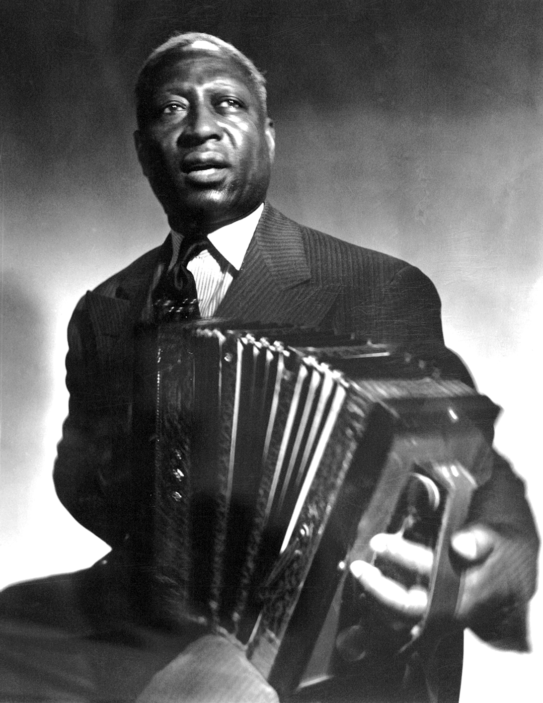 A black and white photograph of a man playing an accordion. The man is African-American and appears to be in his late 60s or early 70s. He is wearing a suit and tie and is holding the accordion in his right hand. He has a serious expression on his face and is looking directly at the camera. The background is blurred so the focus is on the man and his instrument. The photograph is taken from a low angle making the man the focal point of the image.