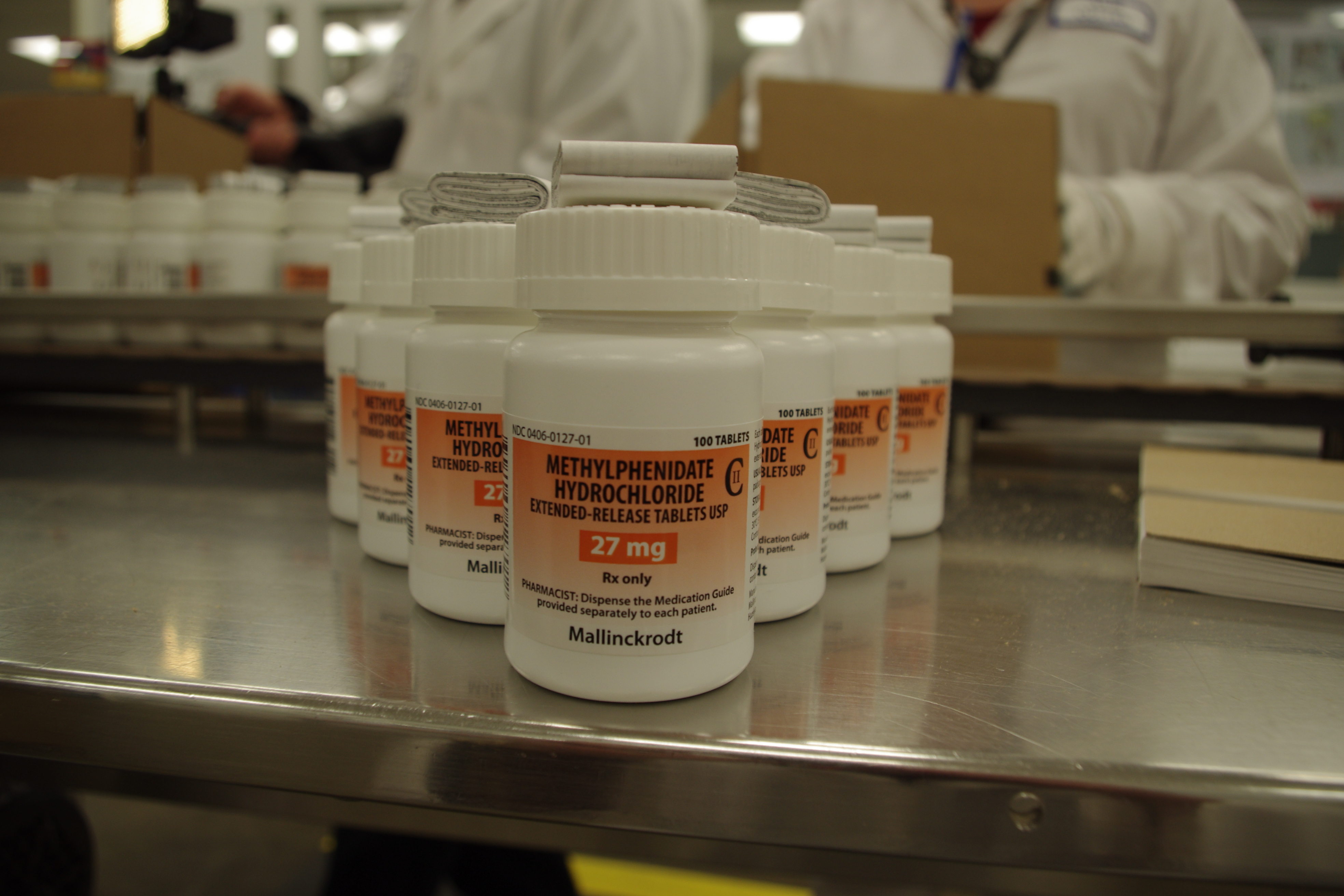 A row of white plastic bottles with orange labels on them. The bottles are arranged on a metal table in a laboratory or laboratory setting. The labels on the bottles read "Methylphenidate Hydrochloride" and "27 mg". There are two people in the background one of whom is holding a cardboard box. The background is blurred but it appears to be a large room with other shelves and equipment.