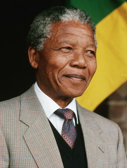 A portrait of Nelson Mandela the former president of South Africa. He is shown in a close-up shot looking directly at the camera with a slight smile on his face. Mandela is wearing a grey suit with a red and blue patterned tie. The background is blurred but it appears to be a brick wall with the South African flag visible behind him.