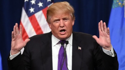 Donald Trump - an American politician media personality and businessman - speaking at a podium while gesturing with both hands. He is wearing a dark suit a white dress shirt with French cuffs cufflinks a purple patterned necktie and an American flag lapel pin. A navy blue curtain the American flag and the European flag are all visible in the background.