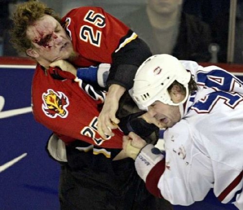 Two ice hockey players engaged in a physical altercation. The player on the left is wearing a red jersey with the number 25 on it and has a bloody face. He is holding onto the other player's arm with both hands and appears to be in the middle of a fight.<br /><br />On the right side of the image there is a player wearing a white jersey with blue and red stripes and a white helmet. He has his mouth open and his eyes are closed indicating that he is in pain or discomfort. The background shows a blue ice rink with spectators in the stands.