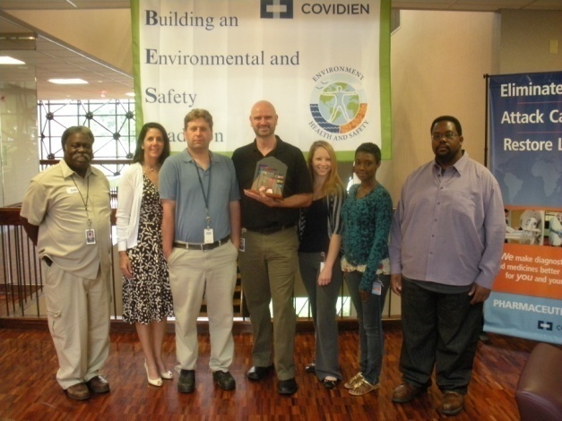 A group of seven people standing in front of a banner that reads "Building an Environmental and Safety" with the logo of Covidien. The banner also has an image of a globe and the text "Eliminate Attack Case Restore Life". The group appears to be posing for a photo in a conference room with a wooden floor and a large window in the background. The people in the photo are of different ages and ethnicities and they are all smiling and looking at the camera.