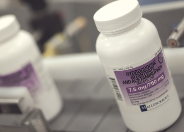 Two white plastic bottles with purple labels on them. The bottles are placed on a gray countertop. The label on the bottles reads "Hydrocodone Bitrate and Acetaminophen 7.5 mg/750 mg". The label also has a barcode and the brand name "Mallinger" written on it. The background is blurred but it appears to be a laboratory setting with other bottles and equipment visible.