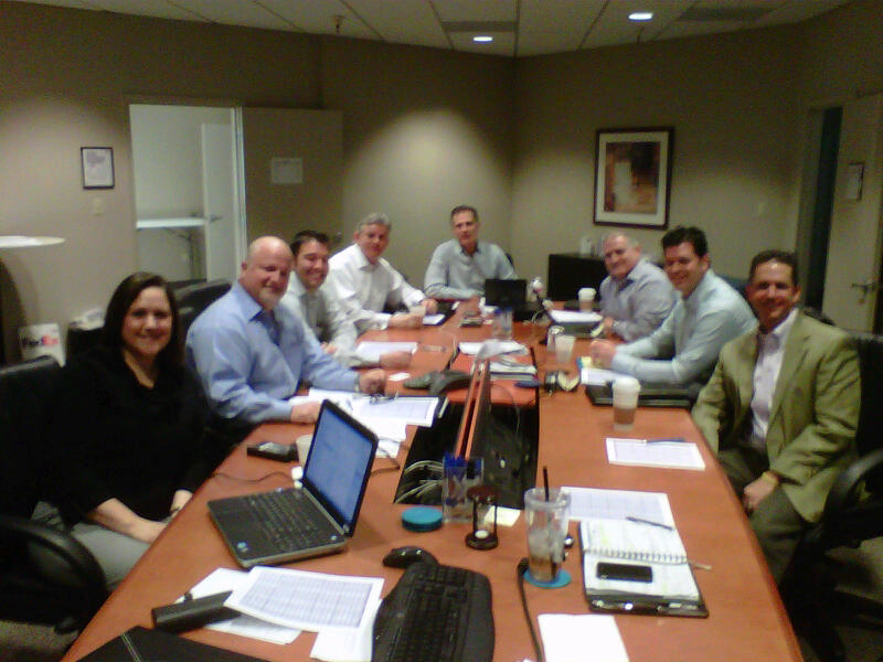 A group of nine people sitting around a long wooden conference table in a meeting room. There are nine people in total nine men and one woman all of whom are smiling and looking at the camera. The table is covered with papers laptops and other office supplies. The room appears to be well-lit with natural light coming in from the windows on the left side of the image. The people are dressed in formal business attire with some wearing suits and others wearing casual clothes. There is a framed picture hanging on the wall in the background.