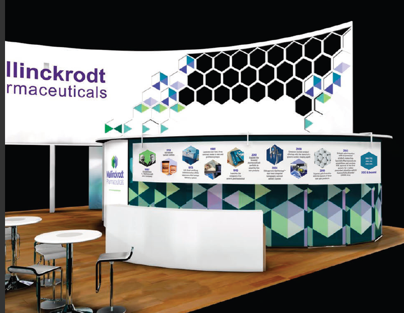 A trade show booth with a white background and a large banner that reads "Mallinckrodt Pharmaceuticals". The booth is decorated with a geometric pattern of blue green and purple hexagons. On the right side of the booth there is a white counter with a blue and green geometric design. Below the counter there are several images and information about the company's products and services. There are also two small white tables and two black barstools in front of the counter. The booth appears to be set up in a room with wooden flooring.