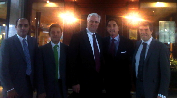 A group of five men standing together in a dimly lit room. They are all dressed in formal attire with suits and ties and appear to be posing for a photo. The man in the center is an older man with white hair and glasses and he is smiling at the camera. The other three men are younger men and they are all looking at the same direction. The room has a wooden floor and a large window in the background and there are plants and a chandelier hanging from the ceiling.