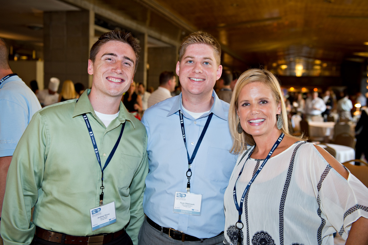 Three people standing in a large room with a high ceiling and chandeliers. They are all smiling and appear to be posing for a photo. The person on the left is wearing a light green button-down shirt and has a name tag around his neck. The man in the middle is in a light blue shirt and is standing next to the woman on the right. The woman is also wearing a white blouse with a black and white pattern. In the background there are other people and tables with white tablecloths.