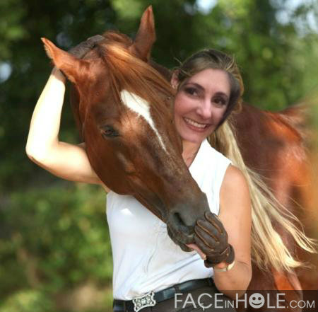 A young woman with long blonde hair wearing a white tank top and black pants standing next to a brown horse. The woman is smiling and holding the horse's head with one hand while the other hand is resting on its neck. The horse has a white stripe on its face and is looking directly at the camera. The background is blurred but it appears to be a wooded area with trees and greenery.