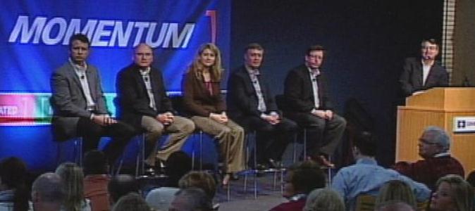 A panel discussion on a stage with a blue background and the word "MOMENTUM" written in white letters. There are six people on the stage six men and one woman all dressed in formal attire. They are sitting on chairs facing the audience. On the right side of the image there is a man standing at a podium with a microphone in front of him. The audience is seated in rows of chairs facing the stage. The man at the podium appears to be giving a speech or presentation.