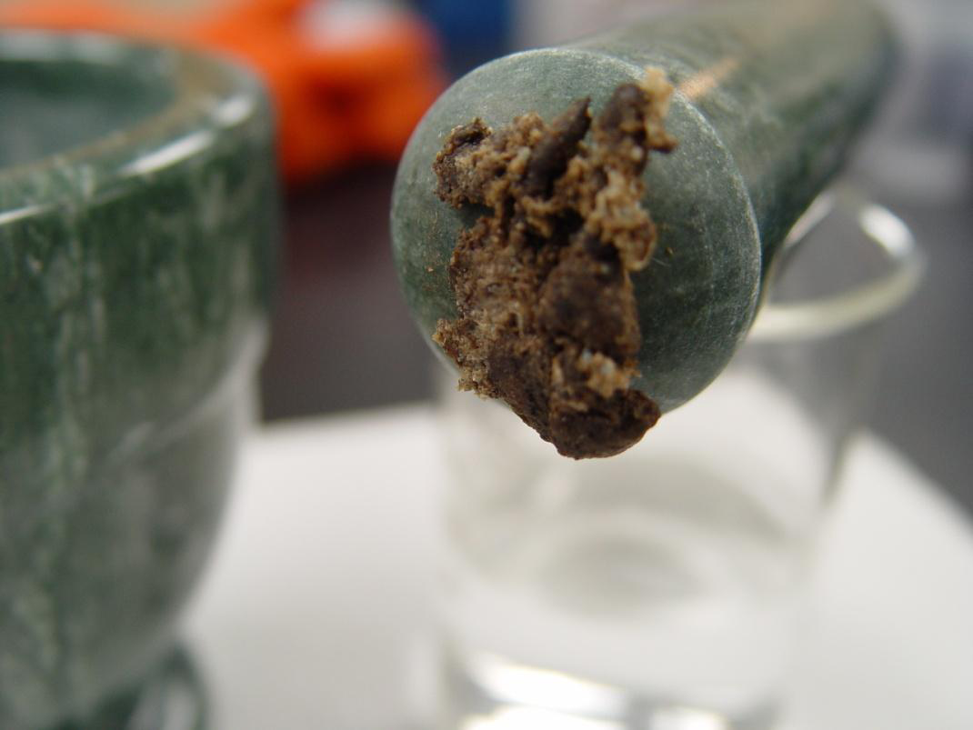 A close-up of a mortar and pestle. The mortar is made of a dark green material possibly stone or marble and has a rough texture. The pestle is resting on top of the mortar and there is a small amount of dirt or grime visible on the surface. The background is blurred but it appears to be a table or countertop with other objects on it.