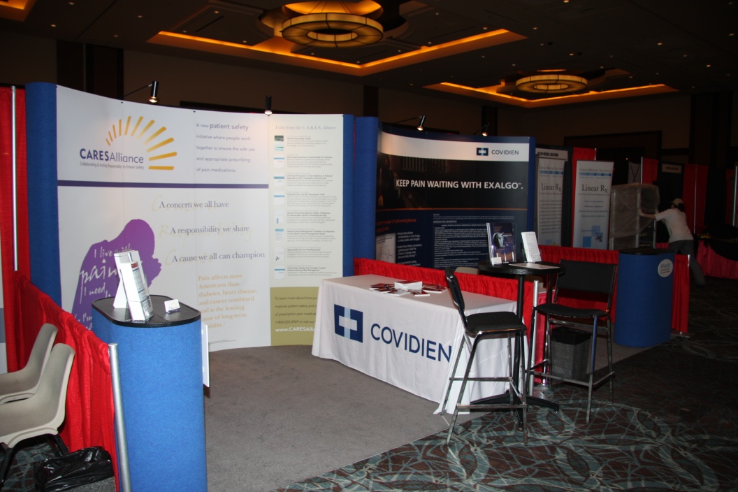 A trade show booth with a blue and white color scheme. The booth is set up in a large room with a carpeted floor and a chandelier hanging from the ceiling. On the left side of the booth there is a large banner with the logo of the company "Covidien" and the company's name in bold letters. Next to the banner there are two smaller banners with the same logo and text. <br /><br />On the right side of this booth it has a table with a white tablecloth and a blue tablecloth with the word "COVIDIEN" written on it. There are two chairs in front of the table and a red curtain in the background.<br /><br />The booth appears to be set up for a conference or event as there are other booths and tables visible in the image. There is also a person standing in the booth and another person walking by.