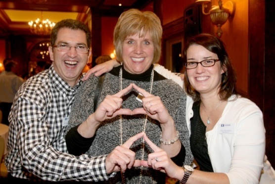 Three people two women and a man posing for a photo in a dimly lit room with chandeliers and other people in the background. The man is on the left side of the image wearing a black and white checkered shirt and glasses and the woman in the middle is in the center. All three are smiling and making a heart shape with their hands. The woman on the right side is wearing a white blouse and glasses. They appear to be happy and enjoying the photo.