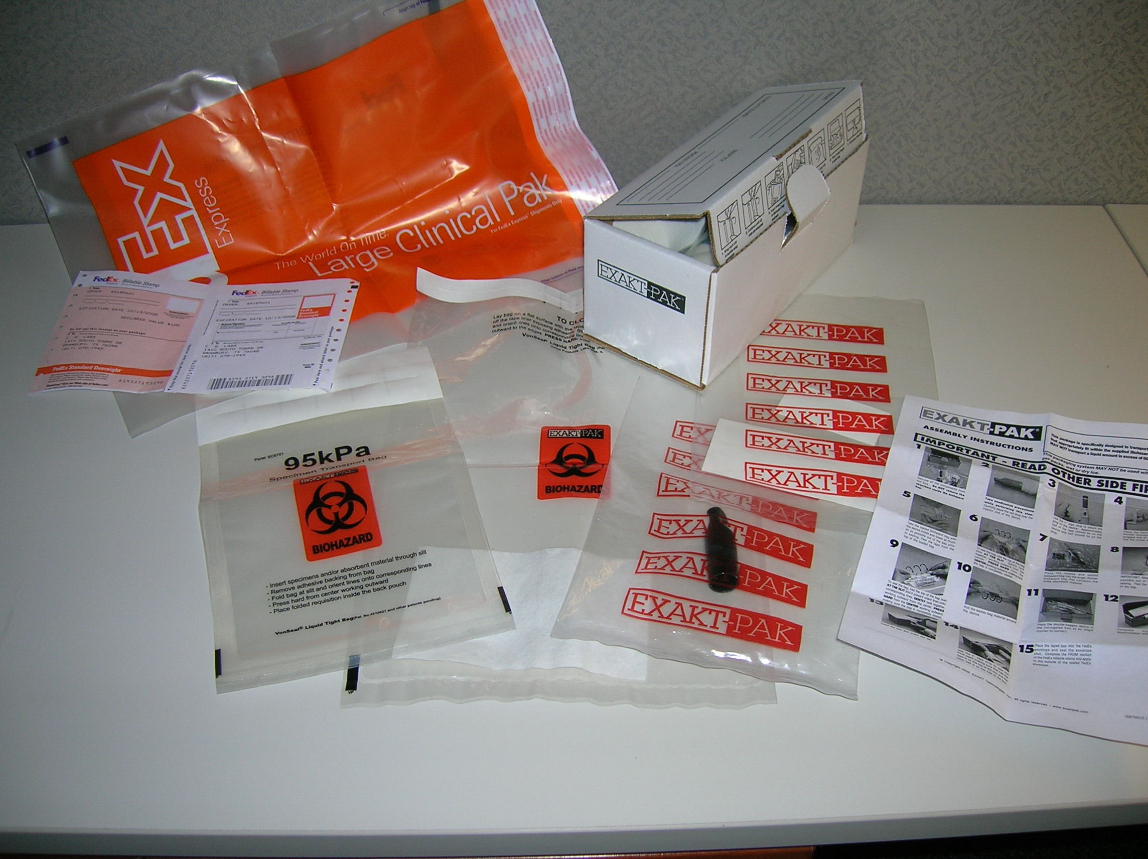 A white table with various items scattered on it. On the left side of the table there is an orange plastic bag with the words "EXPLORER CLINICAL PACK" written on it in white letters. Next to the bag there are two small white boxes with the same words written on them. <br /><br />On the right side of this table on the bottom right corner there appears to be a small white box with a black and white image of a person's face on the front. In the center of the image on top of the white box is a clear plastic bag that has a red and black logo of a biohazard symbol on it.<br /><br />There are also a few other items on the table such as a small piece of paper with instructions on how to use the bag. The background is a plain white wall.