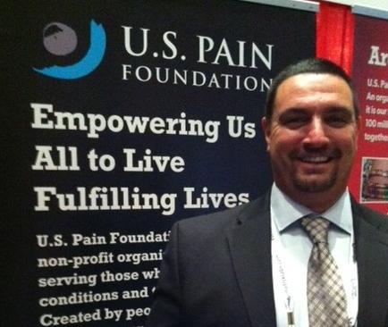 A man standing in front of a banner that reads "U.S. Pain Foundation". The man is wearing a suit and tie and is smiling at the camera. He appears to be in a professional setting possibly at an event or conference. The banner behind him has the logo of the organization and the words "Empowering Us All to Live Fulfilling Lives" in bold white letters. There is also a red banner on the right side of the image with the organization's name and logo.